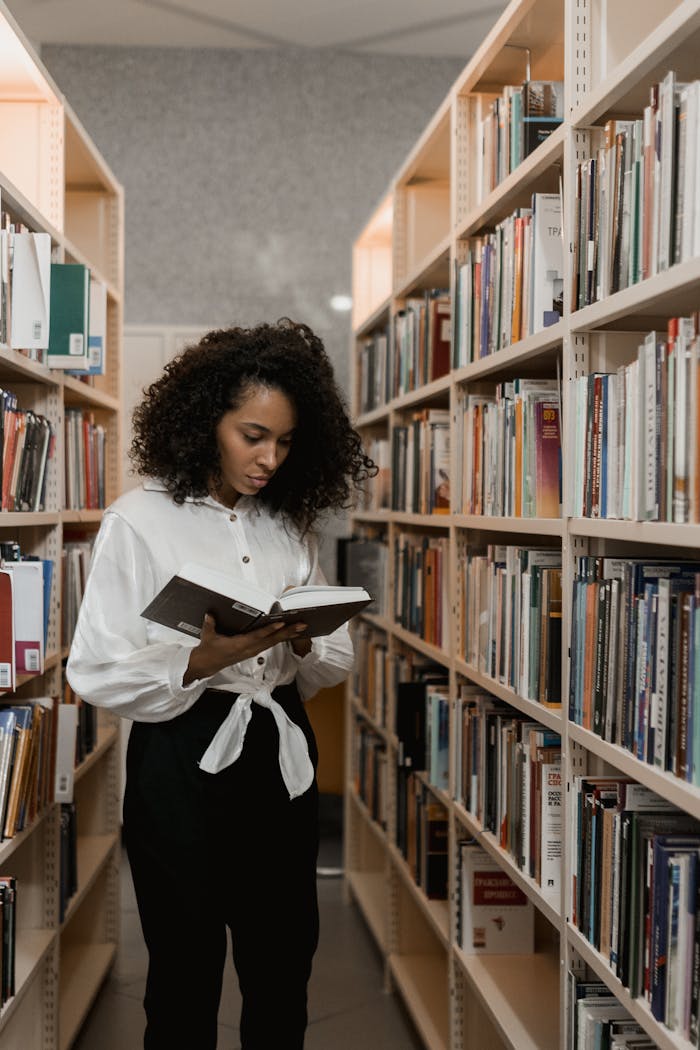 team-02 Focused young woman reading a book in a cozy library aisle surrounded by bookshelves.